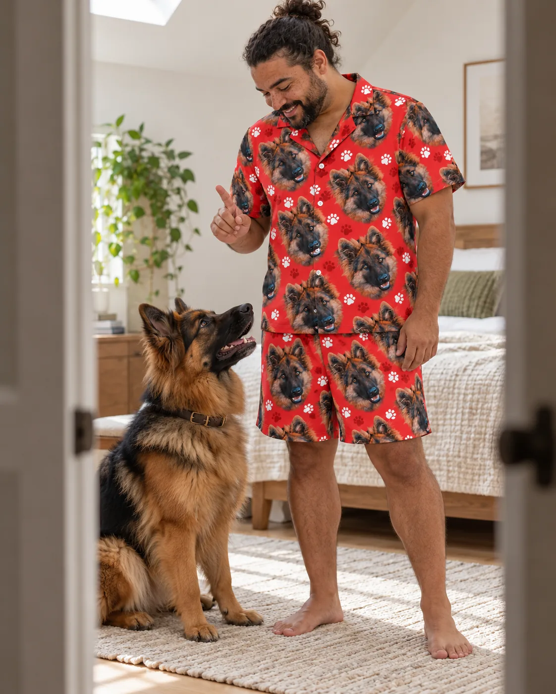 Man in a bright bedroom giving a sit cue to his German shepherd, wearing a red paw print German shepherd pajama set
