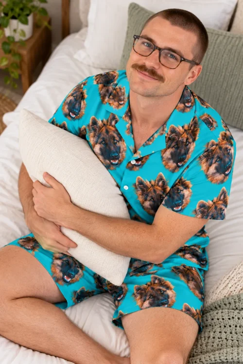 Man in glasses hugging a pillow on a white bed in a teal short-sleeve pajama set with German shepherd face print