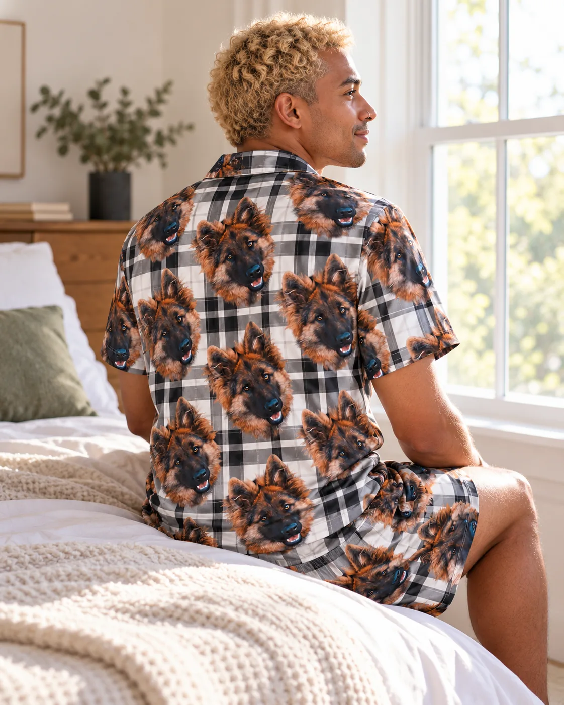 Back view of man sitting on a bed by a sunny window in a white plaid short pajama set with German shepherd faces
