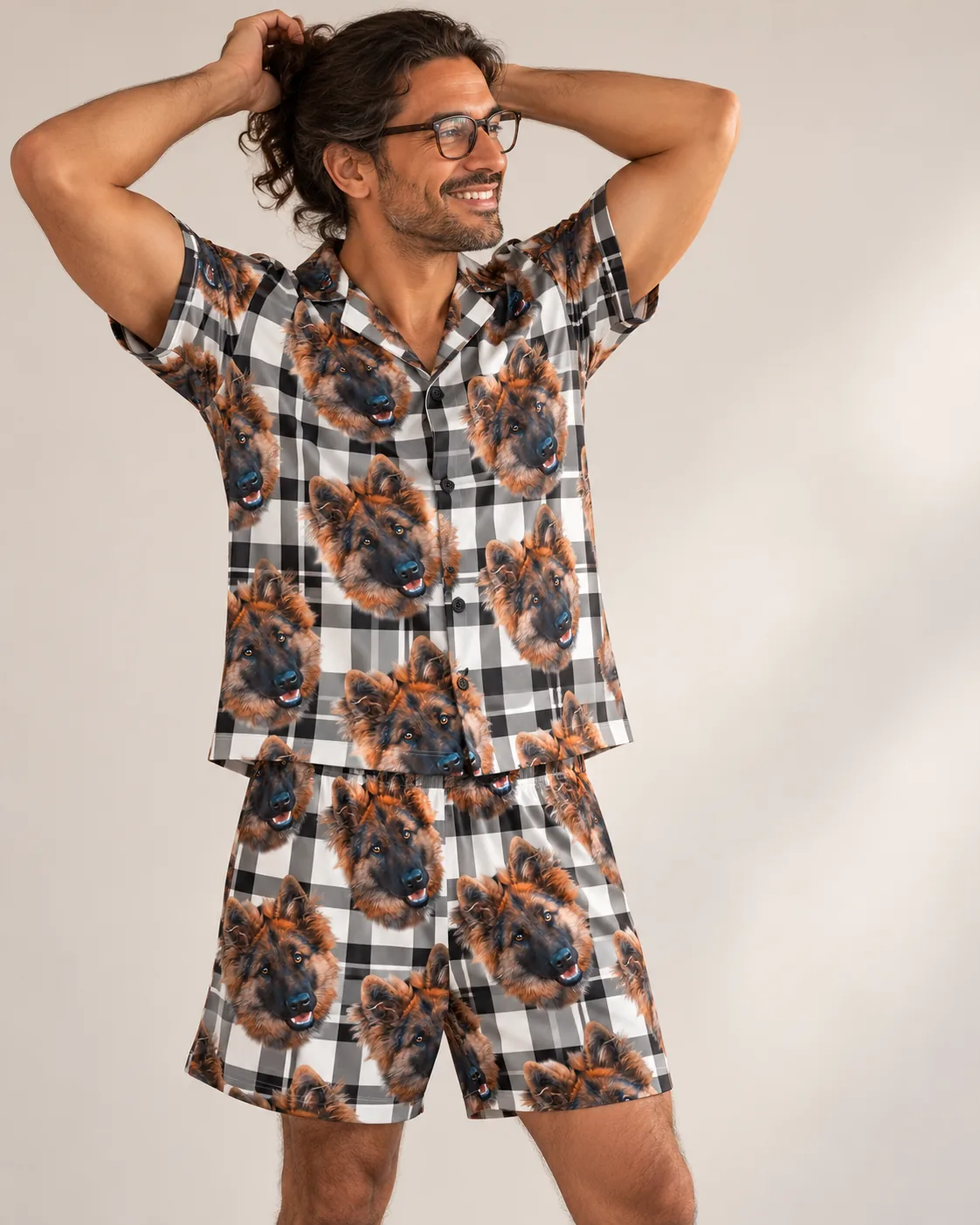 Smiling man in glasses, hands behind head, in a white plaid short pajama set with German shepherd faces, light studio