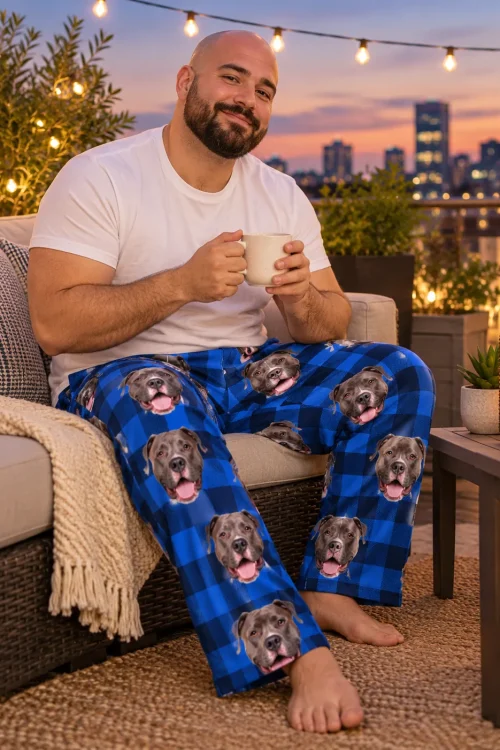 Man with a mug on a rooftop sofa at dusk, city lights behind him, in blue plaid pitbull pajamas