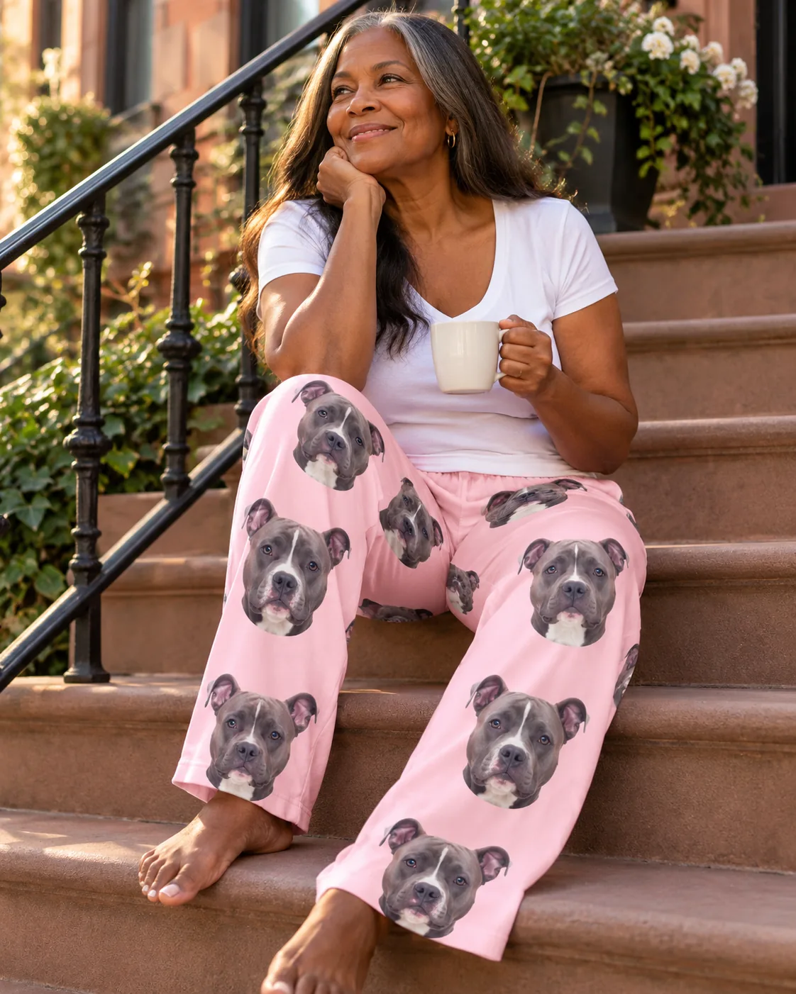 Woman sitting on brownstone steps with a mug, smiling in blush pink pittie pajama pants