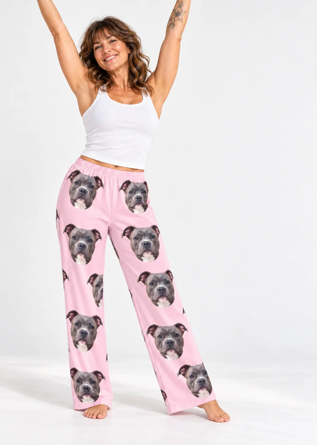 Smiling woman with arms raised in a white tank and blush pink pittie pajamas, barefoot in a white studio