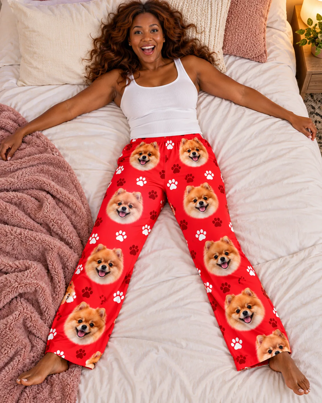 Woman lying arms wide on a bed in red paws pomeranian pajama pants, pink blanket beside her
