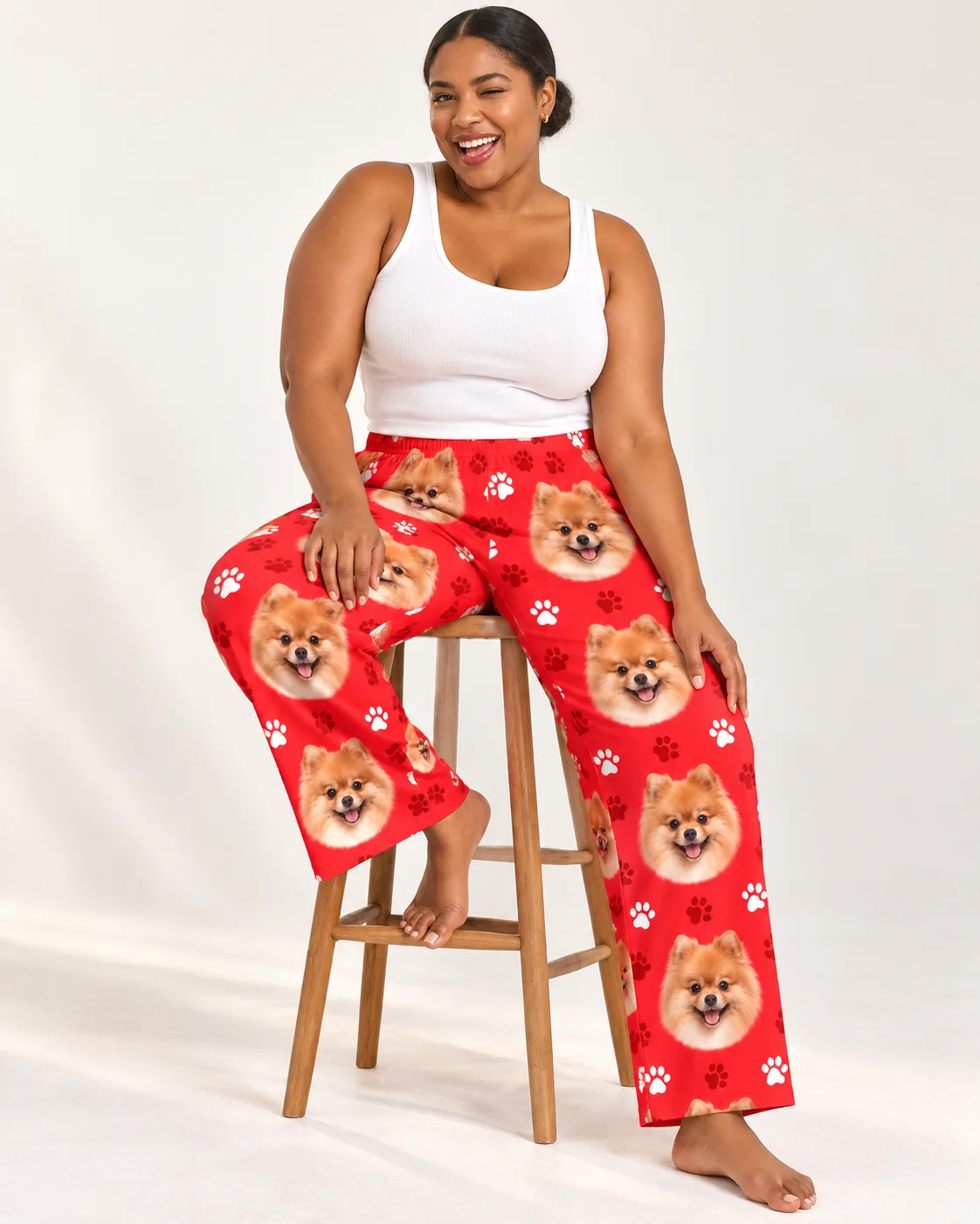 Laughing woman sitting on a wooden stool in red paws pomeranian pajama pants, white tank, cream studio
