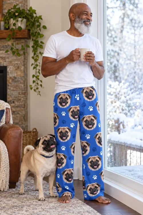 Older man with a mug by a snowy window, his pug beside him, in royal blue paws pug pajama pants
