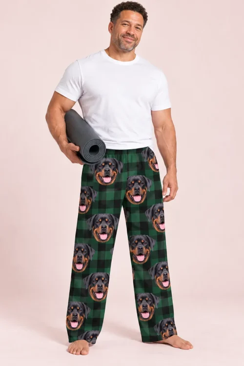 Man holding a rolled yoga mat in green plaid pajama pants with smiling Rottweiler faces, soft pink studio backdrop