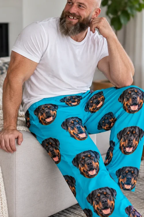 Close-up of teal pajama pants with an elastic waist and smiling Rottweiler faces, worn by a bearded man on a sofa