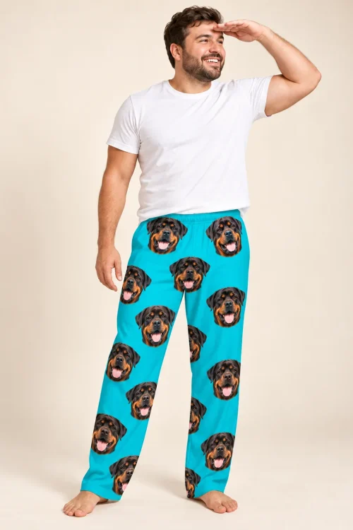 Smiling bearded man shading his eyes in teal pajama pants printed with black and tan Rottweiler faces, cream studio
