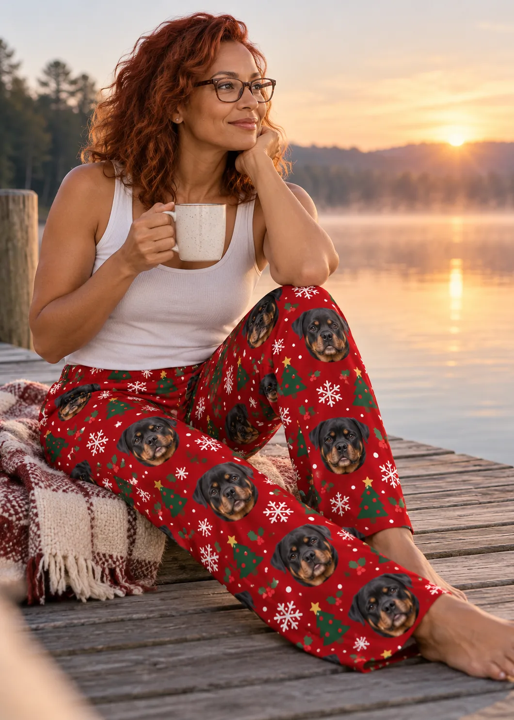 Woman with a mug on a lakeside dock at sunrise in red Christmas pajama pants printed with Rottweiler faces