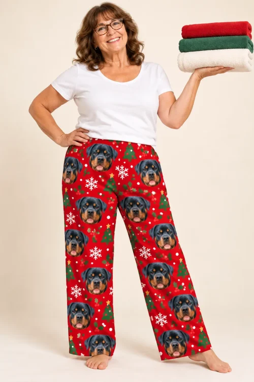 Smiling woman holding folded towels in red Christmas pajama pants with Rottweiler faces and trees, cream studio