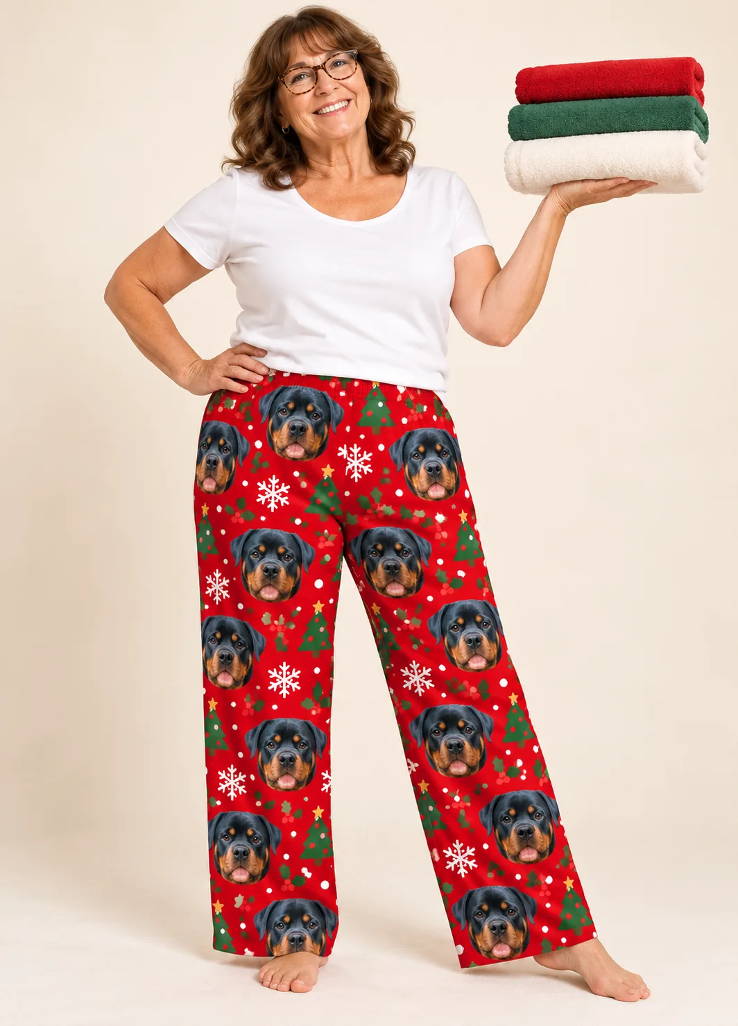 Custom Rottweiler Pajama Pants for Women — christmas studio | GenzOutfit Smiling woman holding folded towels in red Christmas pajama pants with Rottweiler faces and trees, cream studio
