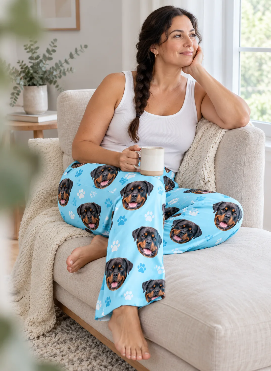 Woman with a mug relaxing on a cream chaise in light blue paw print pajama pants covered in Rottweiler faces