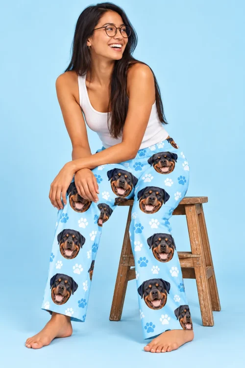 Smiling woman in glasses on a wooden stool in light blue paw print pajama pants with Rottweiler faces, sky blue studio