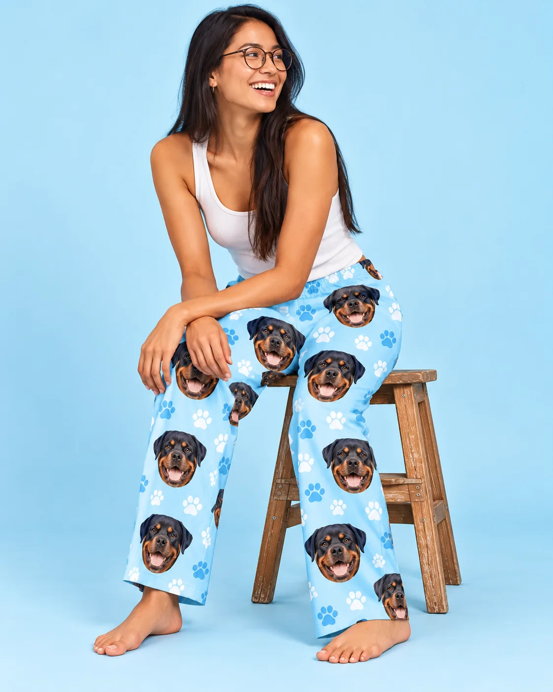 Smiling woman in glasses on a wooden stool in light blue paw print pajama pants with Rottweiler faces, sky blue studio