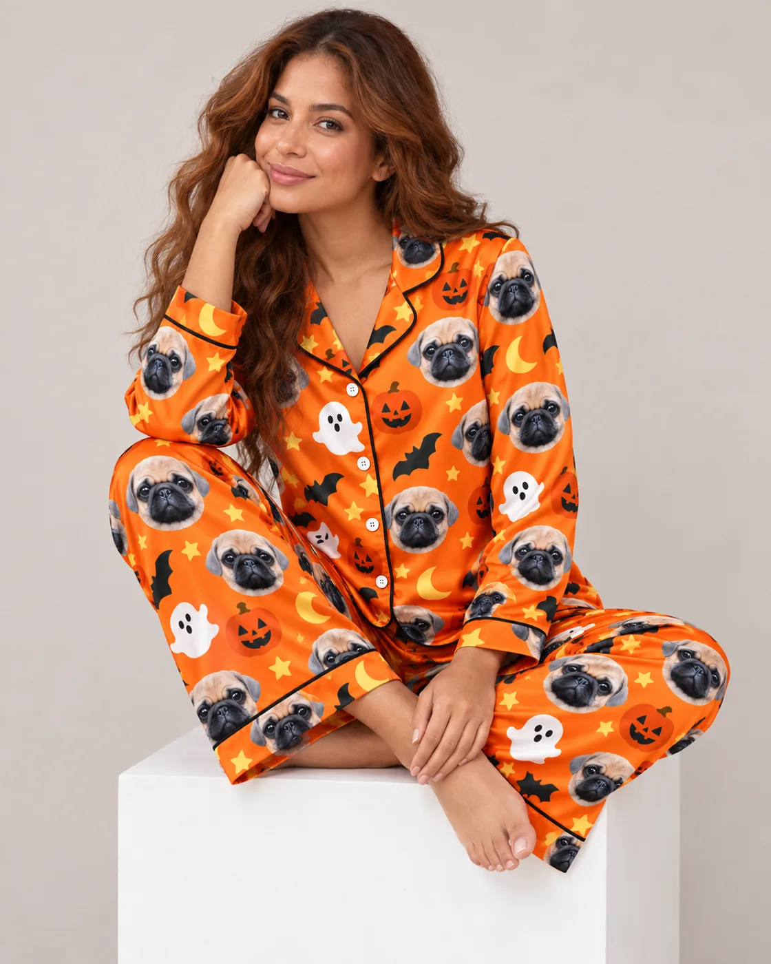 Satin pajamas for women: woman in an orange Halloween satin set printed with pug faces sitting on a white cube