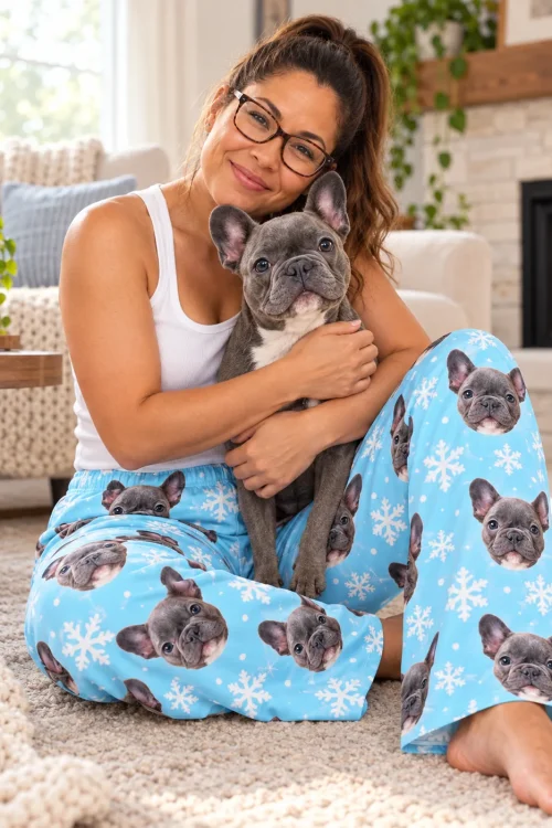 Woman hugging a French bulldog by the fireplace, wearing snowflake pajama pants with its face