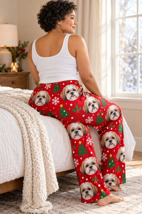 Woman seen from behind, sitting on a bed edge by a snowy window in red Christmas shih tzu pajama pants